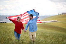 A Couple In Love Having Good Time Walking Through A Meadow With Unfurled American Flag Above Heads. Election, Campaign, Freedom Concept