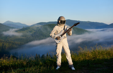 Wall Mural - Astronaut in white space suit and helmet playing guitar, standing on sunny green mountain glade in summer, morning fog rising up from the valley behind him. Concept of astronautics, music and nature.