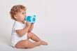 © sementsova321 - Portrait of cute toddler drinking water from bottle while sitting against white wall, wearing body suit, one year old kid with curly hair being thirsty, side view of toddler.