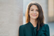 © muse studio - Confident smart woman teacher management student brunette of European appearance stands on the street smiling in a green dress and looks at the camera. Sunny day business center
