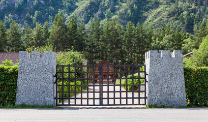 Stylized entrance to the recreation center through the castle gate with stone columns and iron bars with green trees and a wooden house under the mountain. Decorations for the garden.