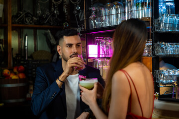 Handsome caucasian man standing at bar counter flirting and talking to beautiful woman with enjoy alcoholic drink cocktail at nightclub party. Smiling couple enjoy romantic dating together at the bar.