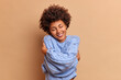 © wayhome.studio - Pleased smiling woman with bushy curly hair embraces herself with love enjoys softness of new jumper feels comfortable and delighted closes eyes in satisfaction isolated over beige background.