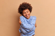 Pleased Smiling Woman With Bushy Curly Hair Embraces Herself With Love Enjoys Softness Of New Jumper Feels Comfortable And Delighted Closes Eyes In Satisfaction Isolated Over Beige Background.