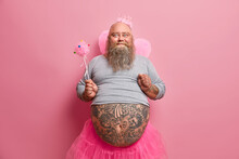 Bearded Adult Man Pretends To Be Priness Makes Your Dreams Come True Looks Positively Wears Wings Undersized Jumper And Pleated Skirt Holds Magic Wand Has Fat Belly With Tattoos. Costume Party
