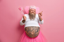 Joyful Bearded Adult Man Fairy Dances Carefree Has Fun On Costume Party Holds Magic Wand Closes Eyes Shows Tattooed Belly Poses Against Rosy Background. People Holiday And Entertainment Concept.