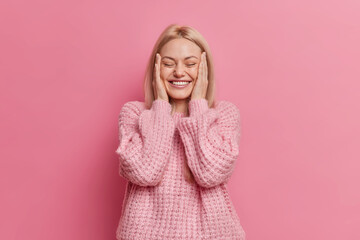 Wall Mural - Positive sincere blonde woman keeps hands on cheeks closes eyes with pleasure dressed in casual winter jumper grins from happiness isolated over rosy studio wall. People and emotions concept