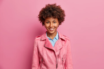 Wall Mural - Stylish pretty dark skinned woman with curly hair pleasant smile dressed in fashionable rosy jacket expresses positive emotions shows optimism poses indoor. Happy emotions and feelings concept