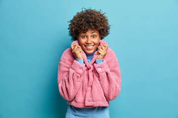 Wall Mural - Portrait of curly haired woman dressed in pink eco coat smiles pleasantly at camera enjoys good day isolated over blue background. Optimistic stylish ethnic lady happy to buy new winter clothes