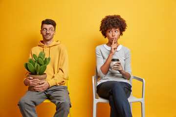 Poster - Mysterious dark skinned woman makes hush gesture and says secret to boyfriend drinks takeaway coffee. Embarrassed guy with pot of cactus smirks face puzzled to hear rumors. Couple on chairs.