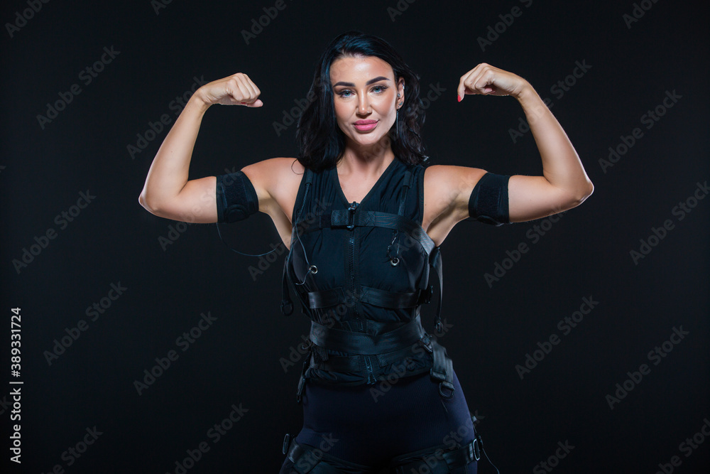 A young athletic woman in an EMS suit on an isolated black background. EMS training. Electro muscular stimulation machine. - obrazy, fototapety, plakaty 