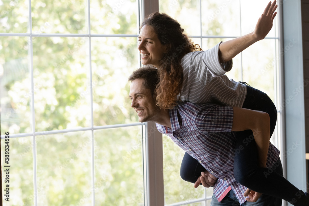 Excited young Caucasian woman piggyback man spouse feel playful relocate to new own house together. Happy millennial couple renters or tenants have fun relax on family leisure weekend at home. - obrazy, fototapety, plakaty 