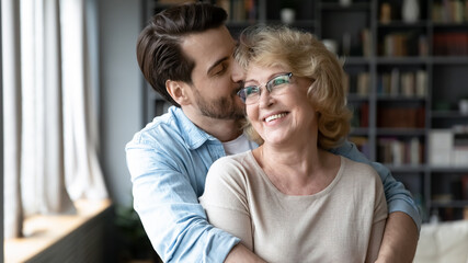 Wall Mural - Loving young handsome man embracing from back and kissing happy older retired mother, showing care and devotion at home. Head shot affectionate grown son feeling appreciation to mature elderly mother.