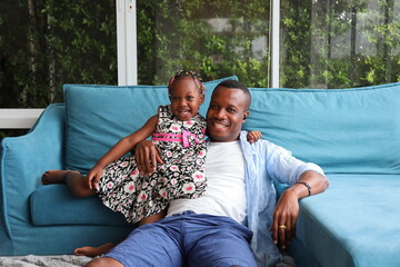 Wall Mural - African American father playing with his little daughter who sitting on the couch while spending happy time together at home on sofa