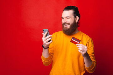 Happy young bearded man looking in his phone and holding a credit card .