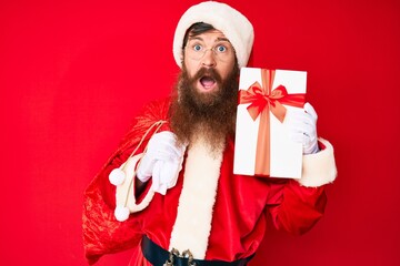 Wall Mural - Handsome young red head man with long beard wearing santa claus costume and big bag with presents clueless and confused expression. doubt concept.