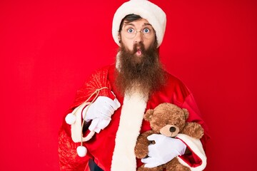 Canvas Print - Handsome young red head man with long beard wearing santa claus costume holding teddy bear making fish face with mouth and squinting eyes, crazy and comical.