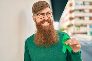 Sticker - Young irish man with redhead beard smiling happy and holding green awareness ribbon leaning on the wall at the city.