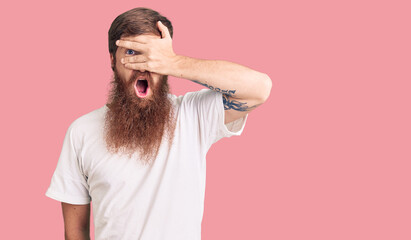 Canvas Print - Handsome young red head man with long beard wearing casual white tshirt peeking in shock covering face and eyes with hand, looking through fingers with embarrassed expression.