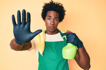 Canvas Print - Handsome african american man with afro hair wearing apron holding cleaning spray with open hand doing stop sign with serious and confident expression, defense gesture