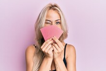 Beautiful Caucasian Blonde Girl Covering Face With Cards Winking Looking At The Camera With Sexy Expression, Cheerful And Happy Face.