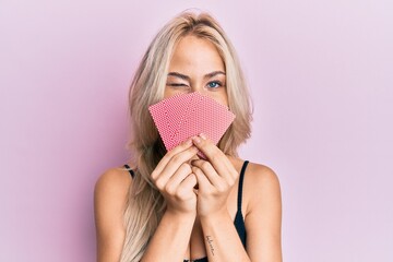 Poster - Beautiful caucasian blonde girl covering face with cards winking looking at the camera with sexy expression, cheerful and happy face.