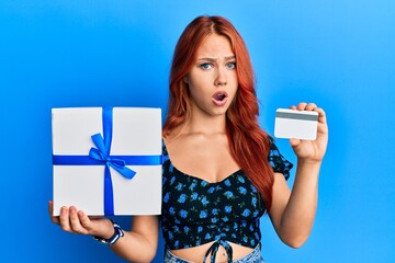 Sticker - Young redhead woman holding gift and credit card in shock face, looking skeptical and sarcastic, surprised with open mouth