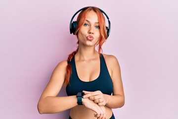 Wall Mural - Young beautiful redhead woman wearing gym clothes, using headphones and smart watch looking at the camera blowing a kiss being lovely and sexy. love expression.