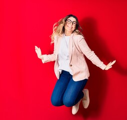 Young beautiful caucasian businesswoman wearing jacket and glasses smiling happy. Jumping with smile on face over isolated red background