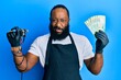 Young african american man tattoo artist wearing professional uniform and gloves holding tattooer machine and russian ruble banknotes clueless and confused expression. doubt concept.