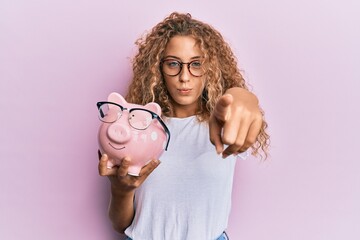 Poster - Beautiful caucasian teenager girl holding piggy bank with glasses pointing with finger to the camera and to you, confident gesture looking serious