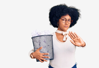 Wall Mural - Young african american girl holding paper bin full of crumpled papers with open hand doing stop sign with serious and confident expression, defense gesture