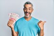 Middle age grey-haired man holding colombian pesos pointing thumb up to the side smiling happy with open mouth