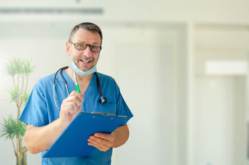 Portrait of a smiling doctor in a blue uniform talking to the camera. The mature man holds a notepad and a pen. There's a stethoscope around his neck.