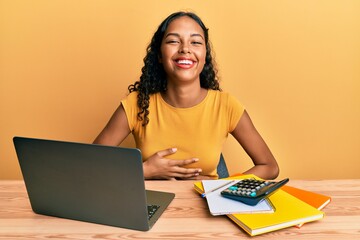 Wall Mural - Young african american girl working at the office with laptop and calculator smiling and laughing hard out loud because funny crazy joke with hands on body.
