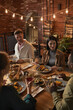 © Seventyfour - Vertical portrait of cheerful adult people sitting at dinner table while enjoying party with outdoor lighting