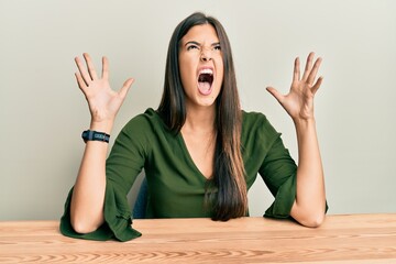 Sticker - Young brunette woman wearing casual clothes sitting on the table crazy and mad shouting and yelling with aggressive expression and arms raised. frustration concept.