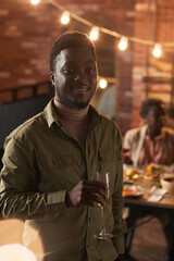 Wall Mural - Vertical waist up portrait of smiling African-American man holding champagne glass and looking at camera while enjoying party outdoor