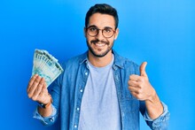 Young Hispanic Man Holding 100 Brazilian Real Banknotes Smiling Happy And Positive, Thumb Up Doing Excellent And Approval Sign