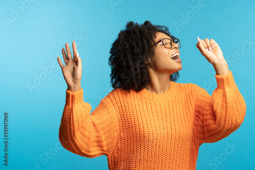 Funny dark skinned millennial woman with curly hair in good mood sings her favorite song, dancing, wear hipster oversize orange sweater, isolated on studio blue background. Mock up, copy space.