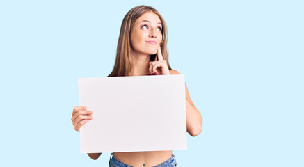 Wall Mural - Young beautiful blonde woman holding blank empty banner serious face thinking about question with hand on chin, thoughtful about confusing idea