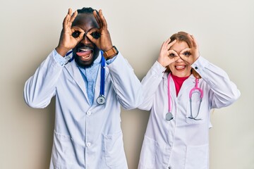 Canvas Print - Young interracial couple wearing doctor uniform and stethoscope doing ok gesture like binoculars sticking tongue out, eyes looking through fingers. crazy expression.