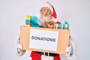 Old senior man wearing santa claus costume holding donation box making fish face with mouth and squinting eyes, crazy and comical.