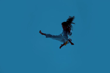 Silhouetted shot of little karate girl in white kimono exercising and fighting, doing martial arts, jumping isolated over blue background