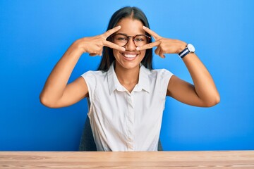 Beautiful hispanic woman wearing casual clothes sitting on the table doing peace symbol with fingers over face, smiling cheerful showing victory