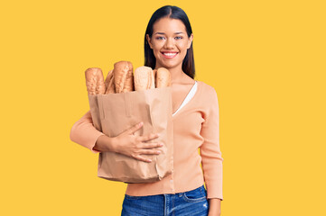 Canvas Print - Young beautiful latin girl holding paper bag with bread looking positive and happy standing and smiling with a confident smile showing teeth