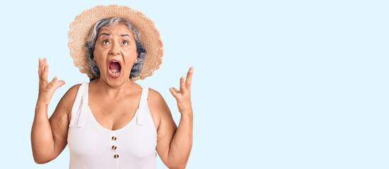 Sticker - Senior woman with gray hair wearing summer hat and summer clothes crazy and mad shouting and yelling with aggressive expression and arms raised. frustration concept.