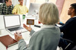 © Svitlana - Close up of aged woman, senior intern holding, taking pills while using laptop, sitting at desk, working in modern office with other young employees
