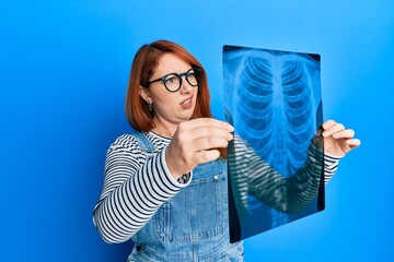 Canvas Print - Beautiful redhead woman holding chest radiography in shock face, looking skeptical and sarcastic, surprised with open mouth