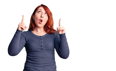 Sticker - Young beautiful redhead woman wearing casual t-shirt amazed and surprised looking up and pointing with fingers and raised arms.